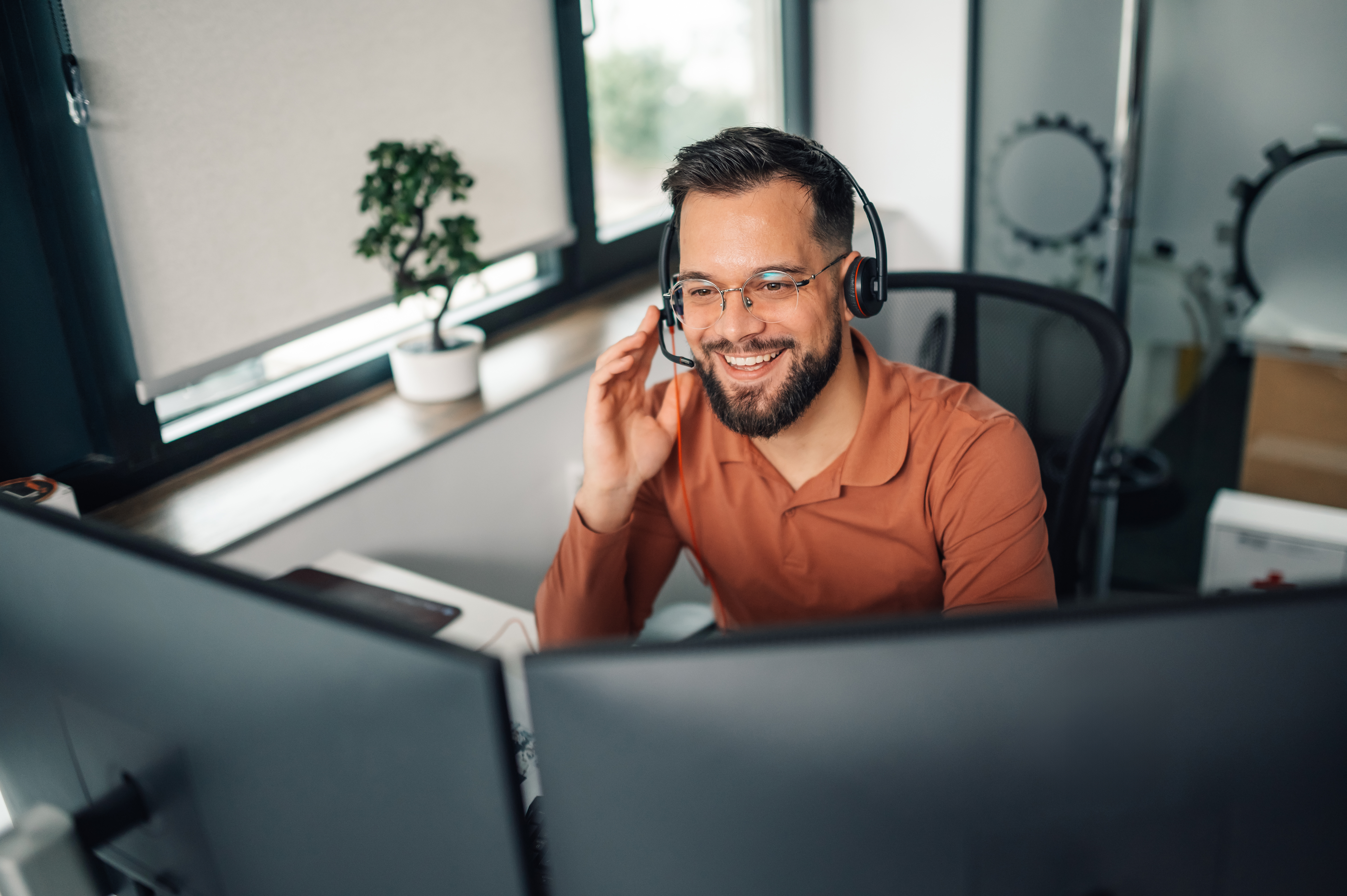 Customer service representative is working from his home office, smiling while using a headset and dual monitors to assist customers or clients