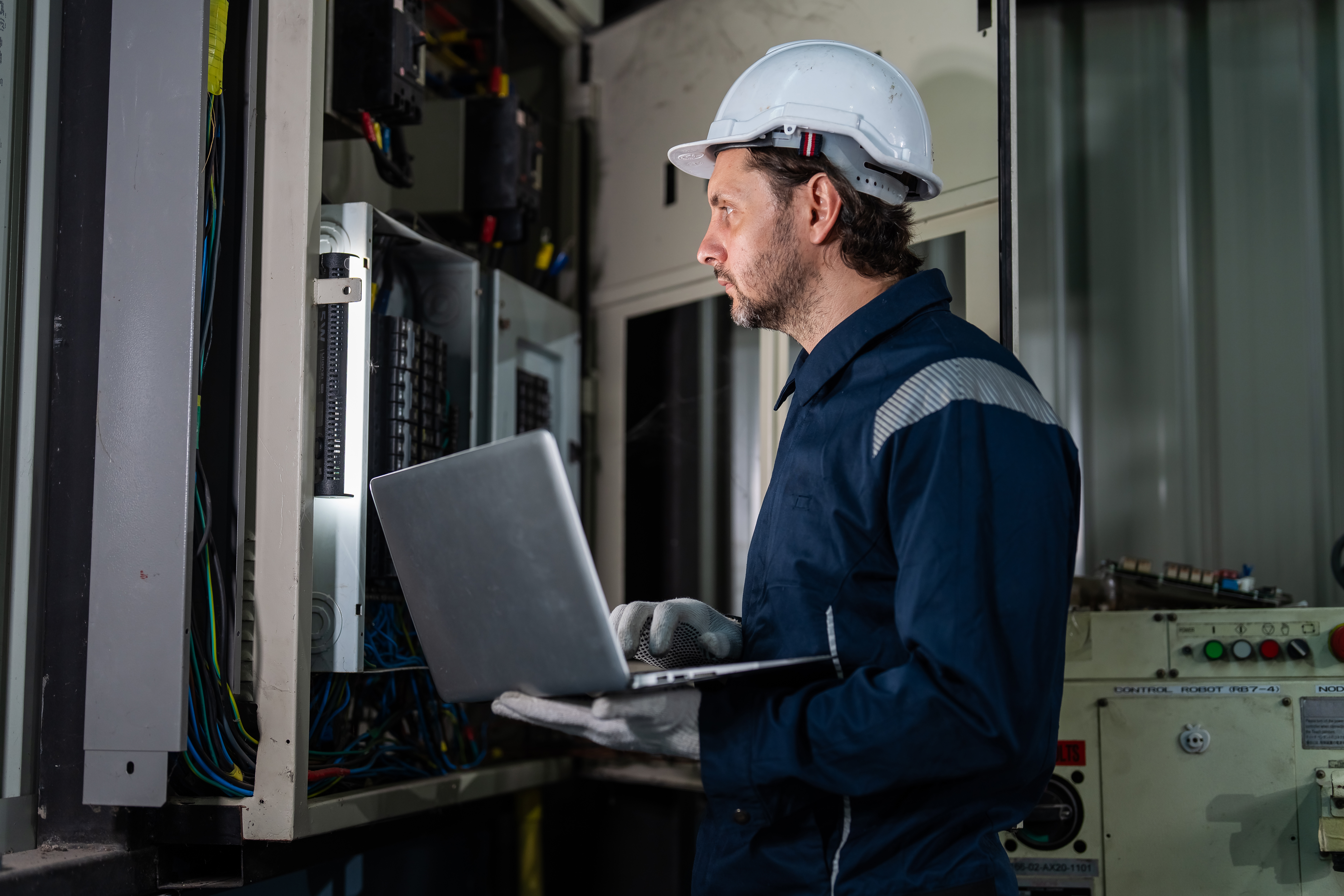 Professional male electrical engineer in uniform using laptop computer for inspecting control panel and switchboard in smart factory. Industrial maintenance and automation technology concept.